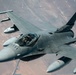 A U.S. Air Force F-16 Fighting Falcon jet fighter is photographed in flight from an aerial perspective, likely from a tanker aircraft during or after aerial refueling. The aircraft is painted in standard gray camouflage and carries multiple external stores on its wing pylons. The terrain visible below appears to be arid desert landscape.