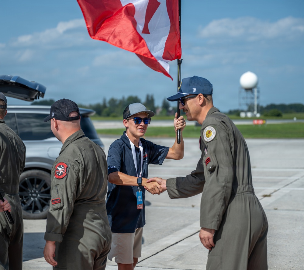 A young civilian wearing a lanyard and holding a Canadian flag shakes hands with a military pilot in an olive flight suit on an airfield tarmac. Several other personnel in flight suits are present nearby. The setting appears to be a military air show or official arrival ceremony.
