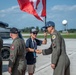 A young civilian wearing a lanyard and holding a Canadian flag shakes hands with a military pilot in an olive flight suit on an airfield tarmac. Several other personnel in flight suits are present nearby. The setting appears to be a military air show or official arrival ceremony.