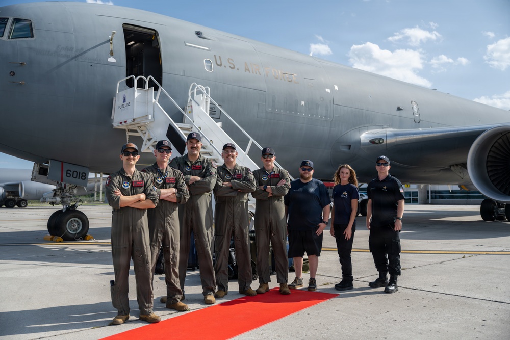 A group of eight personnel pose in front of a U.S. Air Force aircraft on a tarmac. Five individuals are wearing olive/sage green flight suits, while three others are in dark civilian or tactical clothing. They are standing on a red carpet in front of what appears to be a KC-46 Pegasus or similar large military tanker/transport aircraft.