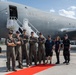 A group of eight personnel pose in front of a U.S. Air Force aircraft on a tarmac. Five individuals are wearing olive/sage green flight suits, while three others are in dark civilian or tactical clothing. They are standing on a red carpet in front of what appears to be a KC-46 Pegasus or similar large military tanker/transport aircraft.