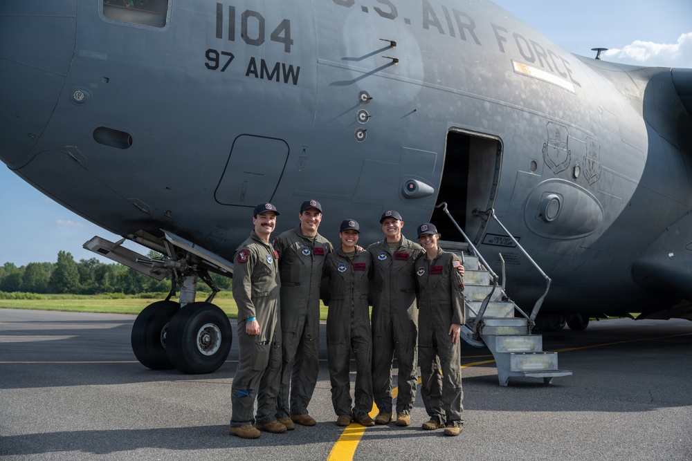 Five U.S. Air Force personnel in flight suits pose together in front of a large military aircraft marked '1104' and '97 AMW' (Air Mobility Wing). The group is standing on a tarmac beneath the fuselage of what appears to be a C-17 Globemaster III, with the aircraft's entry stairs visible to the right. The image appears to be an official or commemorative crew photo.