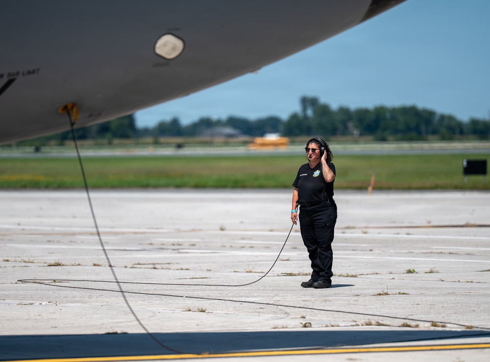 A ground crew member in all-black uniform stands on an airport tarmac beneath the fuselage of a large aircraft. She is wearing a headset and holding a communication cord, likely in contact with the flight crew during pre-flight or post-flight operations. The setting appears to be a military or civilian airfield on a clear, sunny day.
