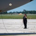 A ground crew member in all-black uniform stands on an airport tarmac beneath the fuselage of a large aircraft. She is wearing a headset and holding a communication cord, likely in contact with the flight crew during pre-flight or post-flight operations. The setting appears to be a military or civilian airfield on a clear, sunny day.