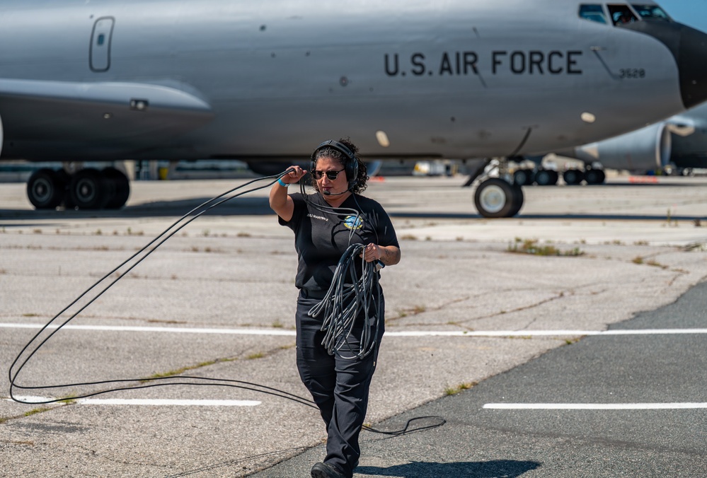 A U.S. Air Force ground crew member walks across a flight line tarmac carrying communication cables and wearing a headset. She is working in front of a large U.S. Air Force aircraft, identifiable by the markings on its fuselage. The setting appears to be a military airfield during daytime operations.