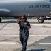 A U.S. Air Force ground crew member walks across a flight line tarmac carrying communication cables and wearing a headset. She is working in front of a large U.S. Air Force aircraft, identifiable by the markings on its fuselage. The setting appears to be a military airfield during daytime operations.