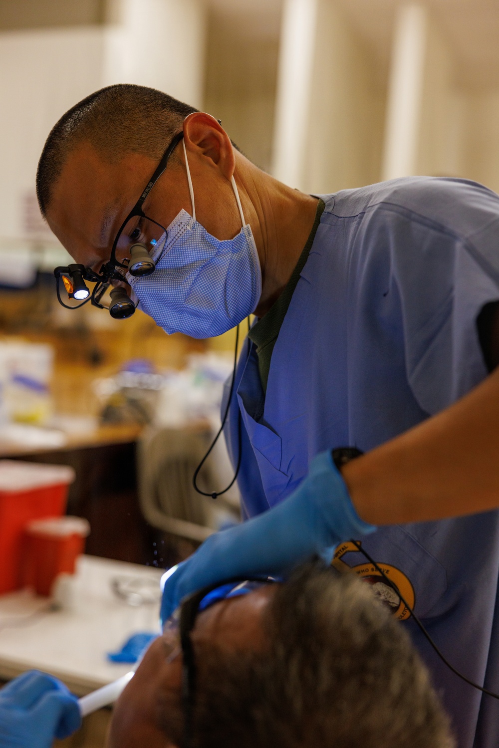 A dental professional wearing blue scrubs, a surgical mask, magnifying loupes with an attached light, and blue nitrile gloves is performing a dental procedure on a patient. The practitioner is leaning over the reclined patient, working inside the patient's mouth. The setting appears to be a dental clinic or field dental station with equipment and supplies visible in the background.