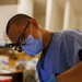 A dental professional wearing blue scrubs, a surgical mask, magnifying loupes with an attached light, and blue nitrile gloves is performing a dental procedure on a patient. The practitioner is leaning over the reclined patient, working inside the patient's mouth. The setting appears to be a dental clinic or field dental station with equipment and supplies visible in the background.