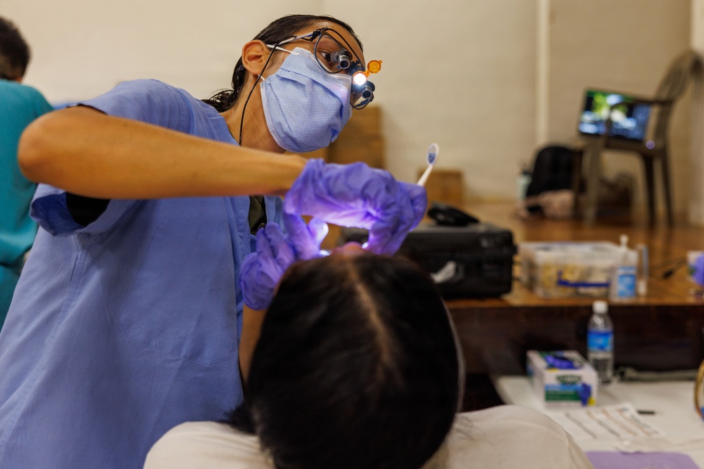 A dental professional wearing blue scrubs, a surgical mask, magnifying loupes with an attached light, and purple nitrile gloves is performing a dental examination or procedure on a reclined patient. The setting appears to be a temporary or community dental clinic, likely a free or outreach event, based on the informal table setup and non-clinical environment. Various dental supplies and equipment are visible on a nearby table.