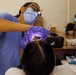 A dental professional wearing blue scrubs, a surgical mask, magnifying loupes with an attached light, and purple nitrile gloves is performing a dental examination or procedure on a reclined patient. The setting appears to be a temporary or community dental clinic, likely a free or outreach event, based on the informal table setup and non-clinical environment. Various dental supplies and equipment are visible on a nearby table.