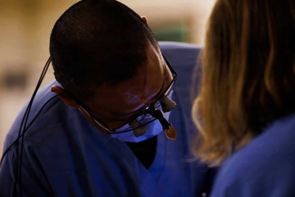 A medical professional wearing surgical loupes (magnification glasses) and a face mask is bent over, focused on a procedure. They are dressed in blue scrubs and appear to be working alongside a second person, also in blue scrubs, visible on the right side of the frame. The image is taken in what appears to be a clinical or surgical setting.