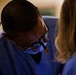 A medical professional wearing surgical loupes (magnification glasses) and a face mask is bent over, focused on a procedure. They are dressed in blue scrubs and appear to be working alongside a second person, also in blue scrubs, visible on the right side of the frame. The image is taken in what appears to be a clinical or surgical setting.