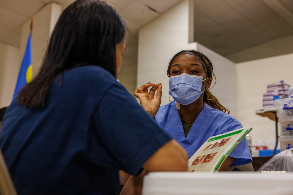A healthcare worker wearing blue scrubs and a light blue surgical mask is engaged in conversation with another person in dark blue scrubs, whose back is to the camera. The setting appears to be a medical or vaccination clinic, with medical supplies and boxes visible in the background. The masked healthcare worker appears to be explaining or consulting, with a pamphlet or informational booklet visible between them.