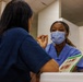 A healthcare worker wearing blue scrubs and a light blue surgical mask is engaged in conversation with another person in dark blue scrubs, whose back is to the camera. The setting appears to be a medical or vaccination clinic, with medical supplies and boxes visible in the background. The masked healthcare worker appears to be explaining or consulting, with a pamphlet or informational booklet visible between them.