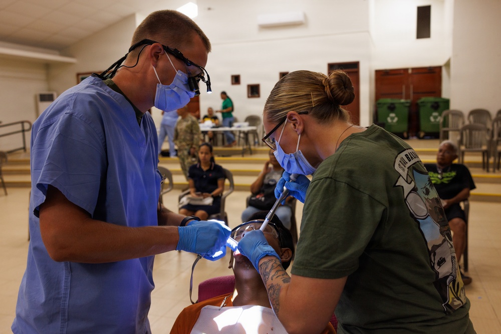 Two medical/dental professionals wearing scrubs, masks, gloves, and protective eyewear are performing a dental procedure on a patient in what appears to be a community hall or public building. The setting suggests a field dental clinic or humanitarian medical mission, with other people visible in the background waiting or assisting. One provider wears blue scrubs with magnification loupes, while the other wears a military-style olive green shirt.