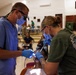Two medical/dental professionals wearing scrubs, masks, gloves, and protective eyewear are performing a dental procedure on a patient in what appears to be a community hall or public building. The setting suggests a field dental clinic or humanitarian medical mission, with other people visible in the background waiting or assisting. One provider wears blue scrubs with magnification loupes, while the other wears a military-style olive green shirt.