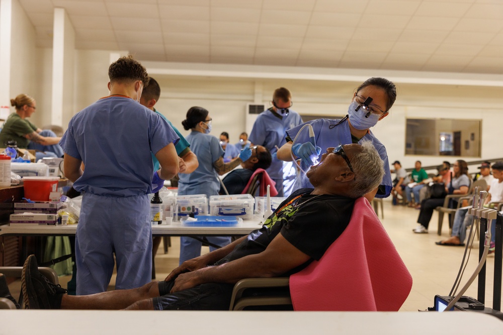 A large-scale community dental clinic is being conducted in what appears to be a gymnasium or community hall. Multiple dental providers in blue scrubs are simultaneously treating patients in portable dental chairs. A waiting area with seated patients is visible in the background, suggesting high demand for services.