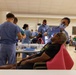 A large-scale community dental clinic is being conducted in what appears to be a gymnasium or community hall. Multiple dental providers in blue scrubs are simultaneously treating patients in portable dental chairs. A waiting area with seated patients is visible in the background, suggesting high demand for services.