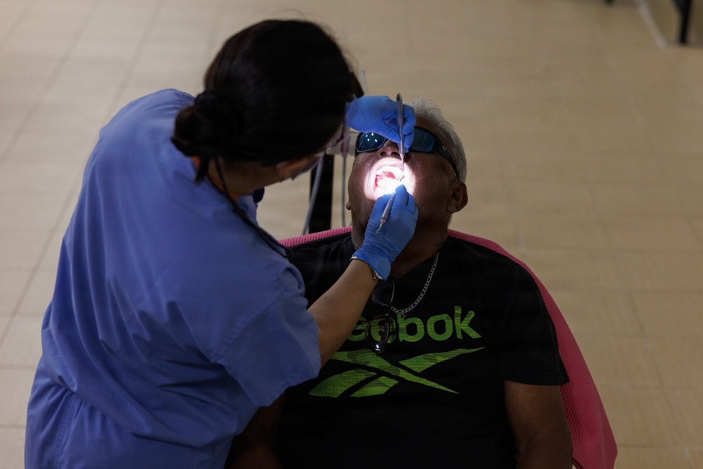A dental professional in blue scrubs and blue latex gloves is performing a dental examination or procedure on a seated patient. The patient, wearing a black Reebok t-shirt and sunglasses, has their mouth open wide while the practitioner uses dental instruments illuminated by a light source. The setting appears to be a non-traditional dental clinic, possibly a community health event or mobile clinic.
