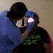 A dental professional in blue scrubs and blue latex gloves is performing a dental examination or procedure on a seated patient. The patient, wearing a black Reebok t-shirt and sunglasses, has their mouth open wide while the practitioner uses dental instruments illuminated by a light source. The setting appears to be a non-traditional dental clinic, possibly a community health event or mobile clinic.