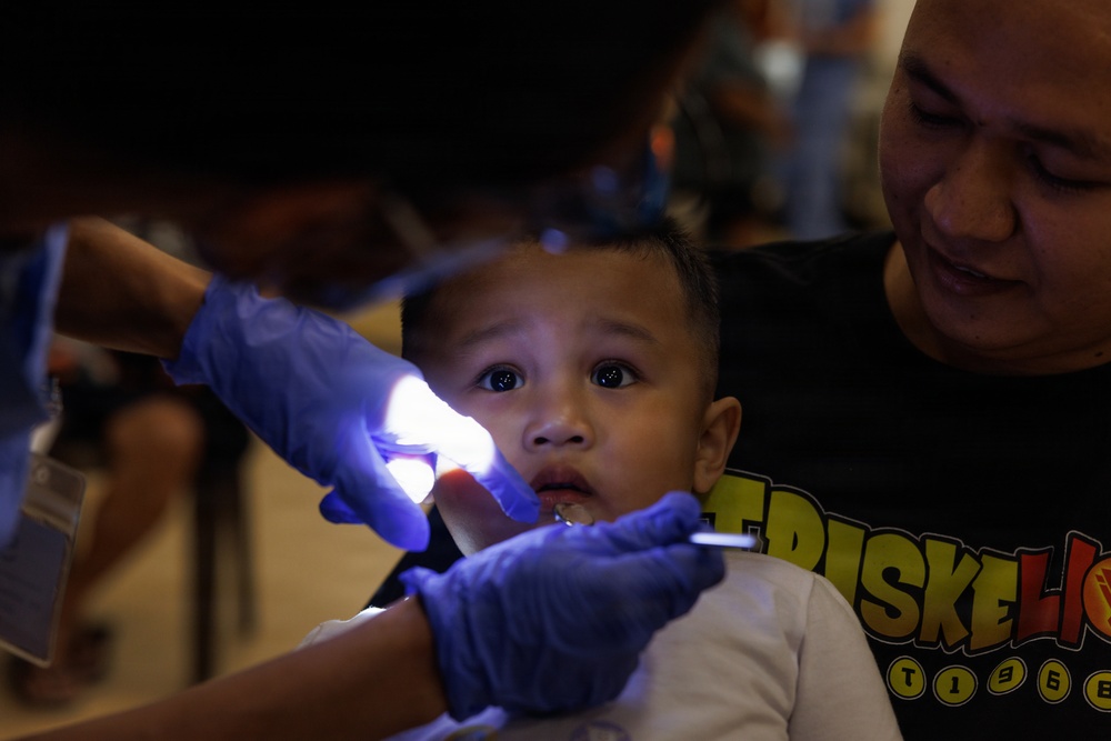 A young toddler is receiving a dental examination while being held by an adult male, likely a parent or guardian. A dental professional wearing blue latex gloves is using a small light and dental instrument to examine the child's mouth. The child appears apprehensive, with wide eyes directed toward the camera.