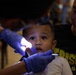 A young toddler is receiving a dental examination while being held by an adult male, likely a parent or guardian. A dental professional wearing blue latex gloves is using a small light and dental instrument to examine the child's mouth. The child appears apprehensive, with wide eyes directed toward the camera.