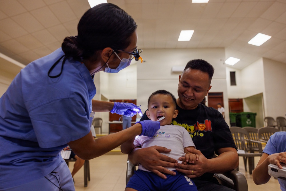 A dental healthcare worker in blue scrubs, surgical mask, and magnifying loupes examines the mouth of a young toddler who is seated on an adult male's lap. The adult male, wearing a black graphic t-shirt, smiles warmly while holding the child securely during the examination. The setting appears to be a community health clinic or outreach event held in a large indoor hall.