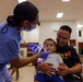 A dental healthcare worker in blue scrubs, surgical mask, and magnifying loupes examines the mouth of a young toddler who is seated on an adult male's lap. The adult male, wearing a black graphic t-shirt, smiles warmly while holding the child securely during the examination. The setting appears to be a community health clinic or outreach event held in a large indoor hall.