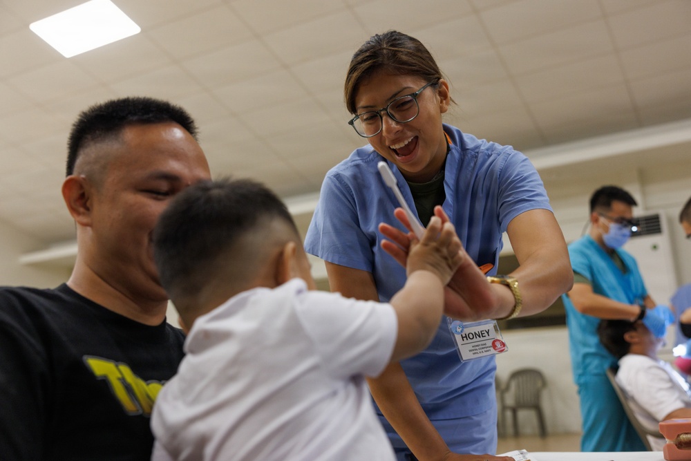 A healthcare worker named 'Honey,' wearing blue scrubs and a name badge, enthusiastically interacts with a young toddler while the child's father looks on smiling. The worker appears to be in a dental or medical outreach setting, holding what looks like a toothbrush or dental instrument while engaging playfully with the child. The scene conveys a warm, positive medical or dental education interaction in what appears to be a community health event.