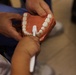 A dental professional in blue scrubs is holding a dental jaw model while a child practices brushing teeth using a small white toothbrush. The scene appears to be a dental education or demonstration session, likely teaching proper brushing technique. The model features realistic-looking white teeth set in an orange/terracotta-colored gum base.