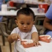 A young child, approximately 2-4 years old, is seated at a white table practicing tooth brushing on a dental jaw model. The child is using a small toothbrush on a red-and-white anatomical teeth model, suggesting a dental hygiene education activity. A second dental model is visible on the table nearby, indicating a structured learning or outreach event.