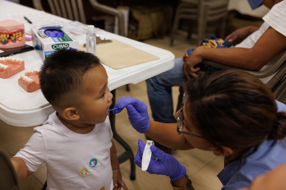 A healthcare or dental professional wearing purple gloves is examining or treating a young child's mouth using a small dental tool. The setting appears to be a community health or dental outreach event, evidenced by dental model props and medical supplies on the table. A second child and adult are visible in the background receiving similar attention.