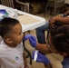 A healthcare or dental professional wearing purple gloves is examining or treating a young child's mouth using a small dental tool. The setting appears to be a community health or dental outreach event, evidenced by dental model props and medical supplies on the table. A second child and adult are visible in the background receiving similar attention.