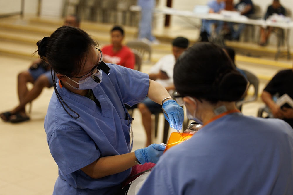 Two dental healthcare workers in blue scrubs, surgical masks, and blue latex gloves are performing a dental procedure on a patient in what appears to be a community health clinic or outreach event. The setting is a large open hall with multiple people waiting in the background. The foreground provider is wearing dental loupes (magnification glasses) mounted on her head.