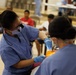 Two dental healthcare workers in blue scrubs, surgical masks, and blue latex gloves are performing a dental procedure on a patient in what appears to be a community health clinic or outreach event. The setting is a large open hall with multiple people waiting in the background. The foreground provider is wearing dental loupes (magnification glasses) mounted on her head.