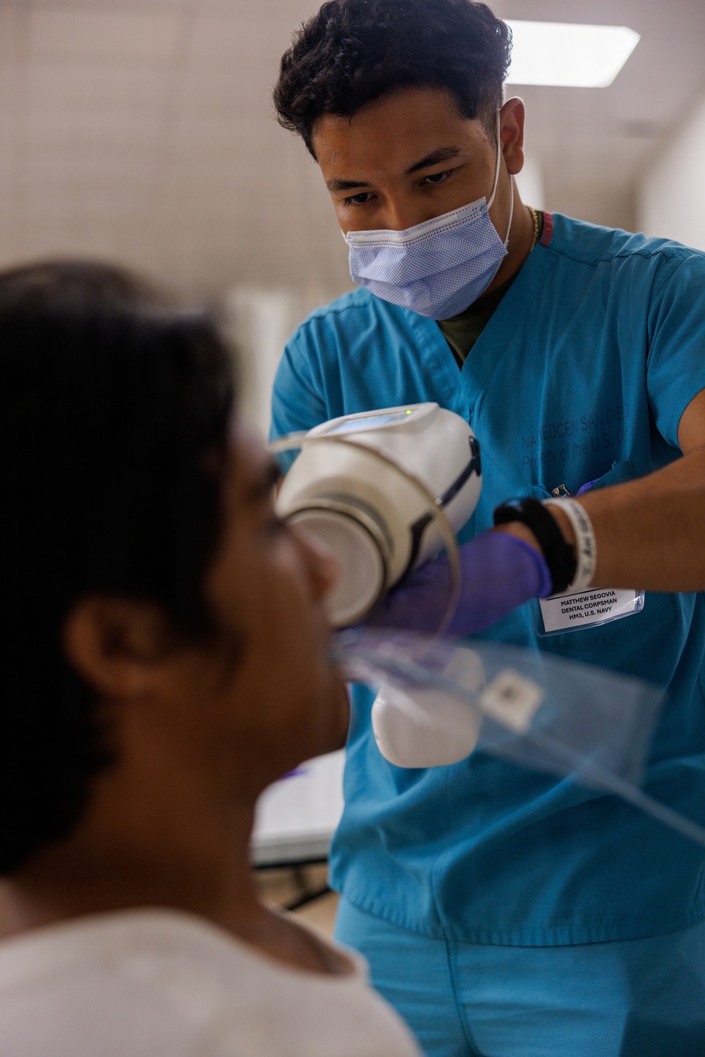 A U.S. Navy Dental Corpsman named Matthew Segovia (HM3) is performing a dental X-ray procedure on a seated patient. He is wearing teal scrubs, a surgical mask, and purple nitrile gloves while positioning an intraoral X-ray device in the patient's mouth. The setting appears to be a clinical dental treatment room.