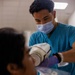A U.S. Navy Dental Corpsman named Matthew Segovia (HM3) is performing a dental X-ray procedure on a seated patient. He is wearing teal scrubs, a surgical mask, and purple nitrile gloves while positioning an intraoral X-ray device in the patient's mouth. The setting appears to be a clinical dental treatment room.