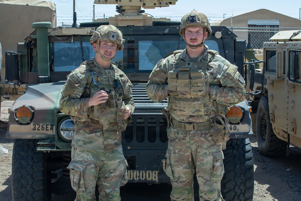 Two U.S. Army soldiers in full combat gear and MultiCam (OCP) uniforms stand in front of a military HMMWV (Humvee) at what appears to be a forward operating base or military installation. Both soldiers are wearing tactical helmets, body armor/plate carriers, and are equipped with various military accessories. The setting appears to be an arid, desert environment based on the terrain and lighting.