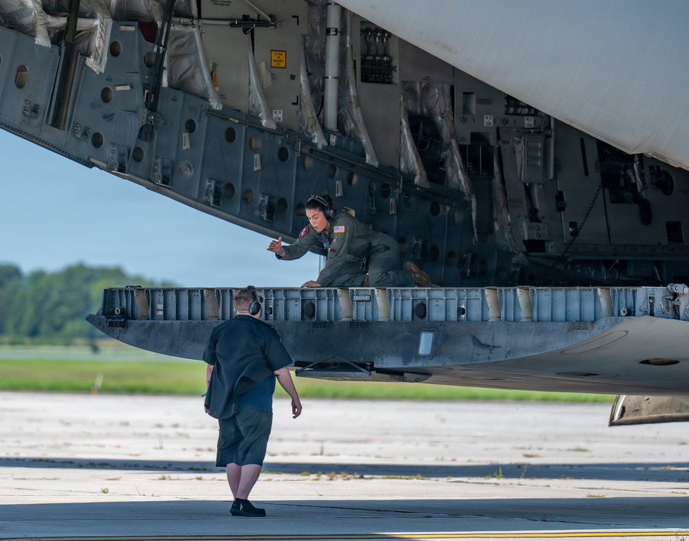 A military aircrew member in an olive green flight suit with a U.S. flag patch is positioned inside the open cargo bay or bomb bay of a large military aircraft, appearing to inspect or operate equipment. A second individual in dark civilian-style clothing and wearing headphones stands on the tarmac below, observing or communicating with the crew member. The scene takes place on a paved airfield with green trees visible in the background.