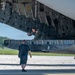 A military aircrew member in an olive green flight suit with a U.S. flag patch is positioned inside the open cargo bay or bomb bay of a large military aircraft, appearing to inspect or operate equipment. A second individual in dark civilian-style clothing and wearing headphones stands on the tarmac below, observing or communicating with the crew member. The scene takes place on a paved airfield with green trees visible in the background.