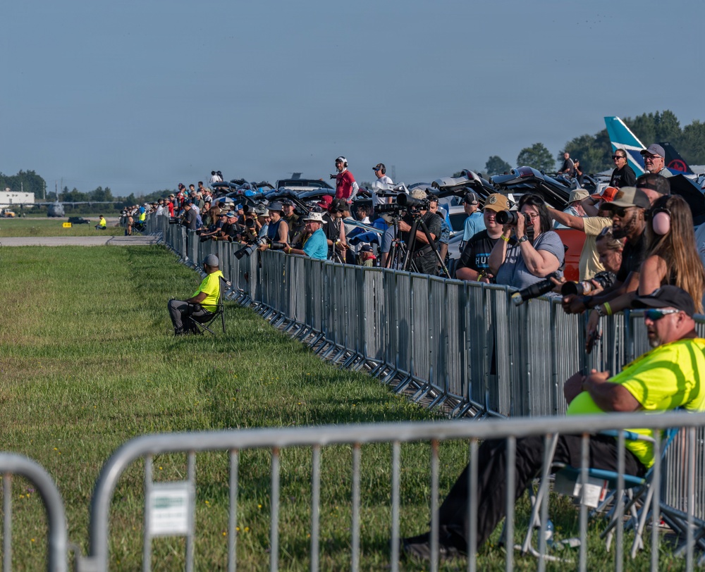 A large crowd of spectators and photographers lines a metal crowd-control barrier at what appears to be an airshow. Many attendees are equipped with cameras, telephoto lenses, and hearing protection, watching aircraft on the tarmac. Staff members in high-visibility yellow vests are visible on the airside of the barrier.