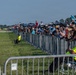 A large crowd of spectators and photographers lines a metal crowd-control barrier at what appears to be an airshow. Many attendees are equipped with cameras, telephoto lenses, and hearing protection, watching aircraft on the tarmac. Staff members in high-visibility yellow vests are visible on the airside of the barrier.