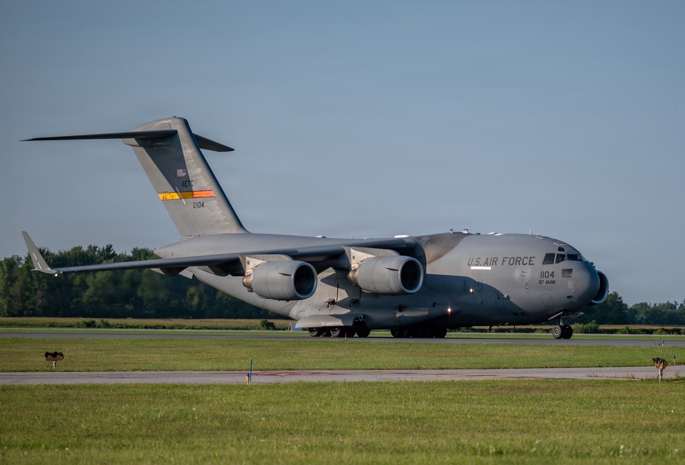 A U.S. Air Force C-17 Globemaster III military cargo aircraft is taxiing on a runway or taxiway. The aircraft bears tail number 21104 and unit designation '97 AMW' (Air Mobility Wing), painted in standard military gray livery. The photo was taken during golden hour lighting conditions at what appears to be a military or civilian airfield.