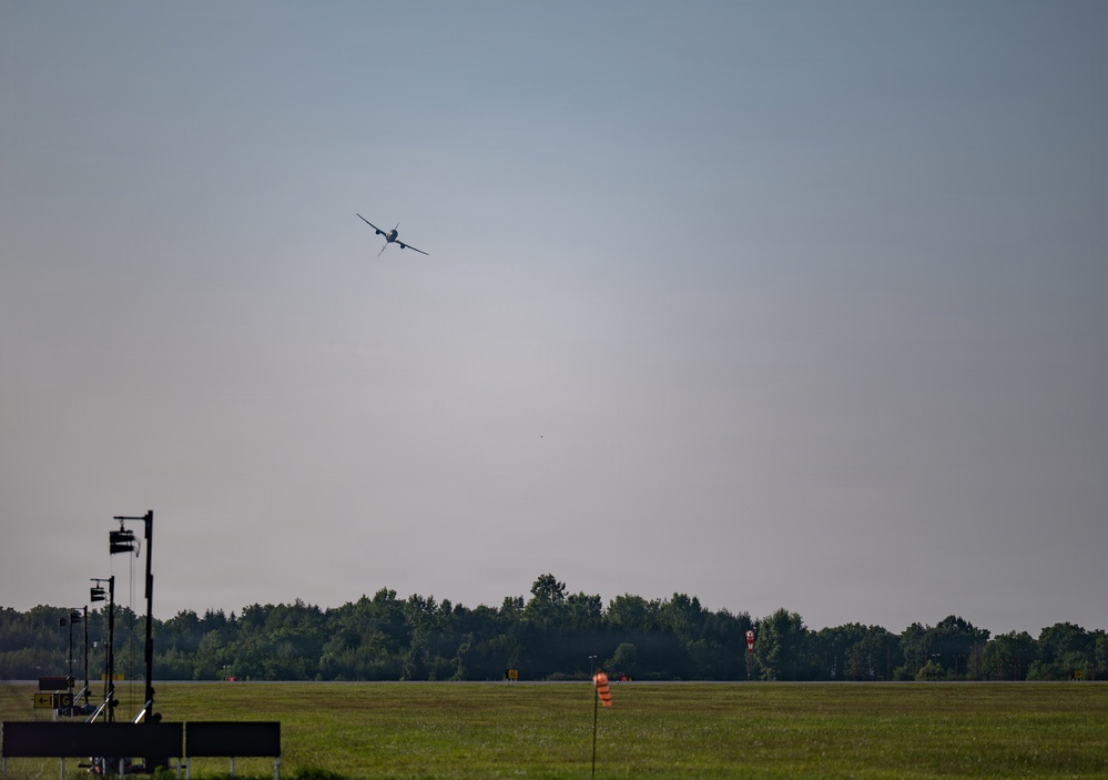 An aircraft is captured in flight at altitude above an airfield, appearing to be in a climbing or banking maneuver. The foreground shows airport infrastructure including approach lighting structures and a windsock on a flat grassy field. A treeline forms the background horizon under a hazy, overcast sky.