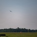 An aircraft is captured in flight at altitude above an airfield, appearing to be in a climbing or banking maneuver. The foreground shows airport infrastructure including approach lighting structures and a windsock on a flat grassy field. A treeline forms the background horizon under a hazy, overcast sky.