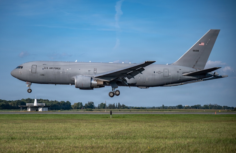 A U.S. Air Force Boeing KC-46A Pegasus tanker aircraft with tail number 66018 is captured on approach to landing with landing gear fully deployed. The aircraft is painted in standard military gray livery and bears U.S. Air Force markings along with an American flag on the tail. The photo is taken at what appears to be a military or civilian airfield with green grass, a treeline, and a small ground structure visible in the background.