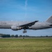 A U.S. Air Force Boeing KC-46A Pegasus tanker aircraft with tail number 66018 is captured on approach to landing with landing gear fully deployed. The aircraft is painted in standard military gray livery and bears U.S. Air Force markings along with an American flag on the tail. The photo is taken at what appears to be a military or civilian airfield with green grass, a treeline, and a small ground structure visible in the background.