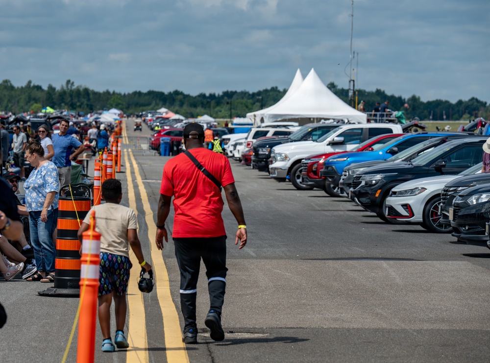 An outdoor automotive event, likely a car show or drag racing event, taking place on a wide paved runway or strip. A man in a red shirt and a child walk along the central lane, flanked by rows of parked vehicles on the right and spectators on the left. Orange traffic cones separate the walking/racing lane from the crowd area, with white event tents visible in the background.