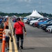An outdoor automotive event, likely a car show or drag racing event, taking place on a wide paved runway or strip. A man in a red shirt and a child walk along the central lane, flanked by rows of parked vehicles on the right and spectators on the left. Orange traffic cones separate the walking/racing lane from the crowd area, with white event tents visible in the background.
