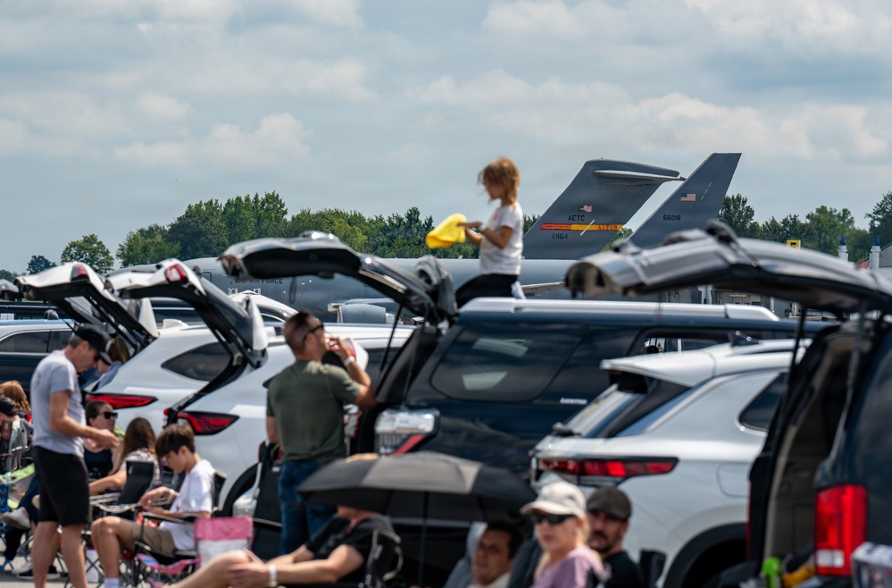 A crowded parking lot at what appears to be a military air show, with spectators gathered around their vehicles with open trunks to watch the event. A military jet aircraft tail fin is visible in the background, marked with 'AETC' and the serial number '66018', indicating it belongs to Air Education and Training Command of the U.S. Air Force. People are seated in folding chairs and standing between vehicles in warm, partly cloudy weather.